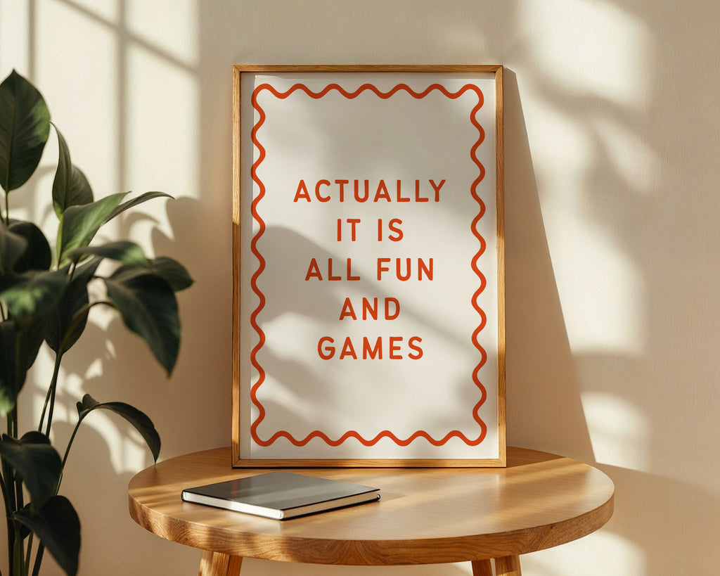 Framed quote 'Actually it is all fun and games' on a wooden table with a plant in the background