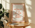 Framed quote 'Actually it is all fun and games' on a wooden table with a plant in the background