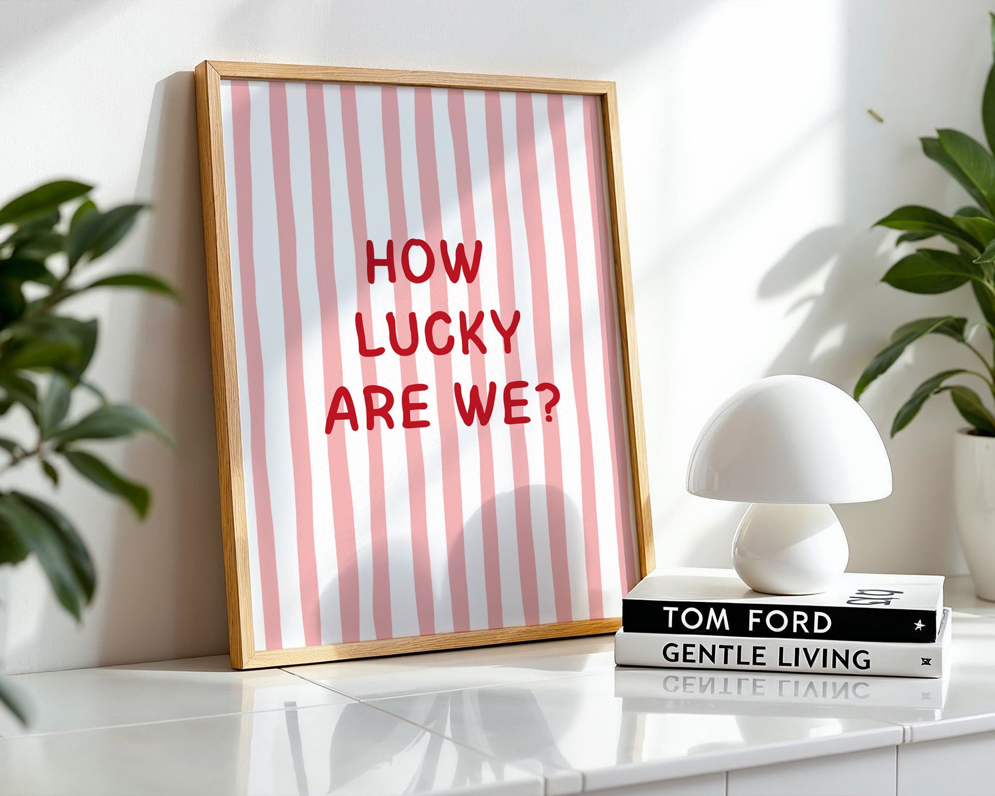 Framed print with 'HOW LUCKY ARE WE?' text on a striped background, placed on a surface with books and a lamp.