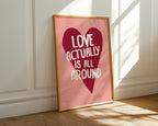 Framed poster with heart design and text 'Love actually is all around' on a pink background, leaning against a white wall.