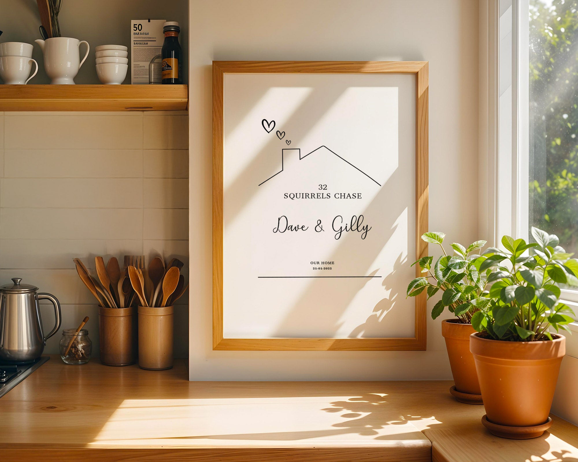 Framed artwork on a kitchen counter with a house design and text, surrounded by plants and kitchen items.