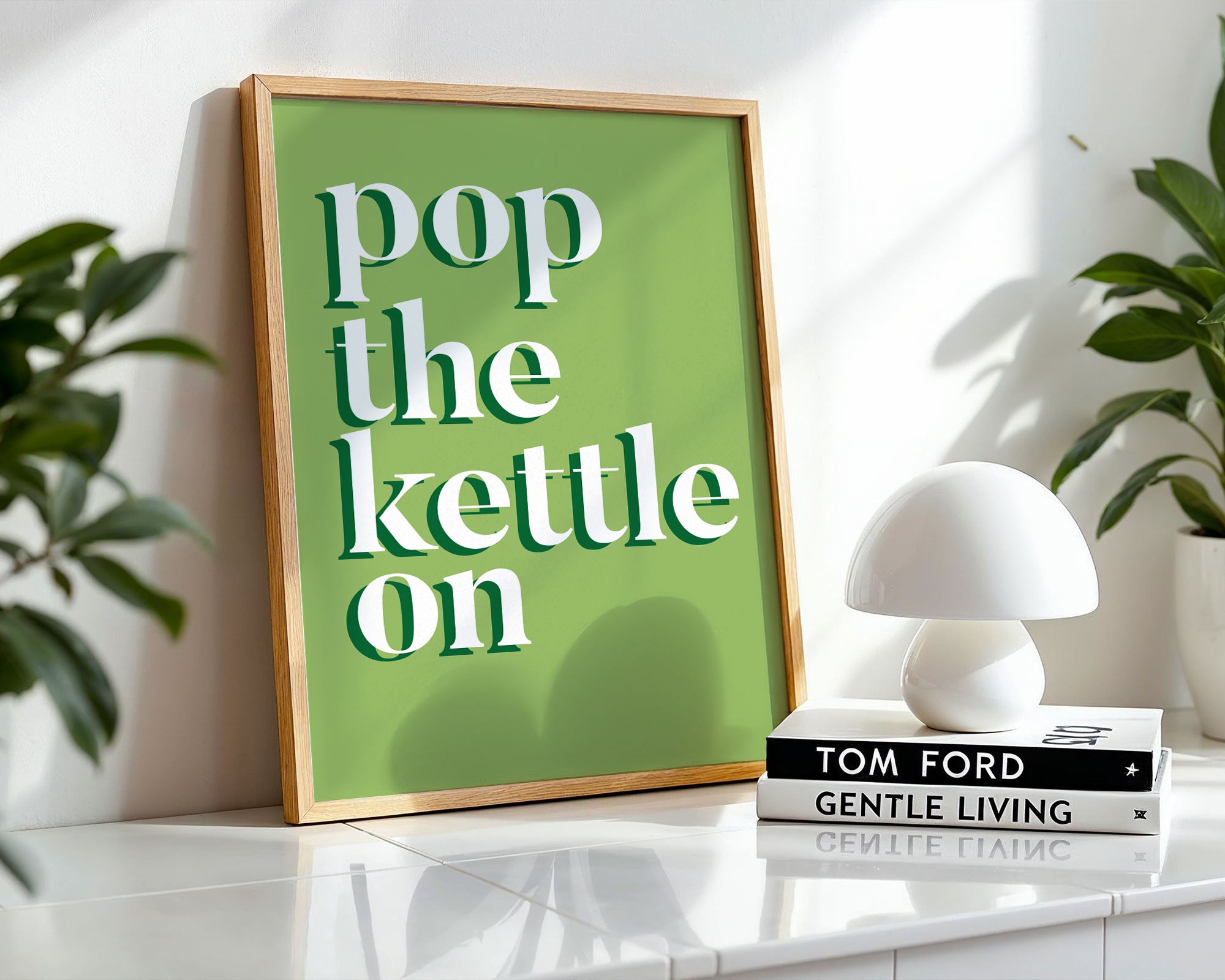Framed poster with 'pop the kettle on' text on a shelf with books and a lamp.