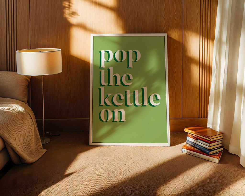 Framed artwork with 'pop the kettle on' text in a cozy room with books and a lamp.