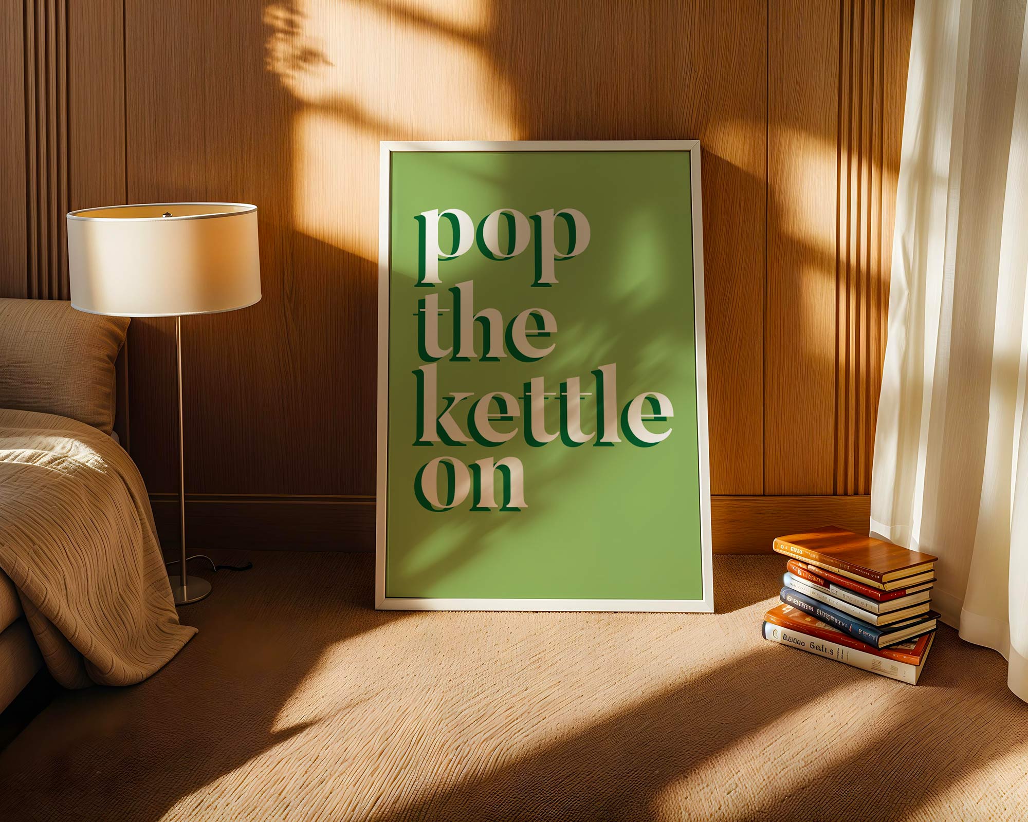 Framed artwork with 'pop the kettle on' text in a cozy room with books and a lamp.