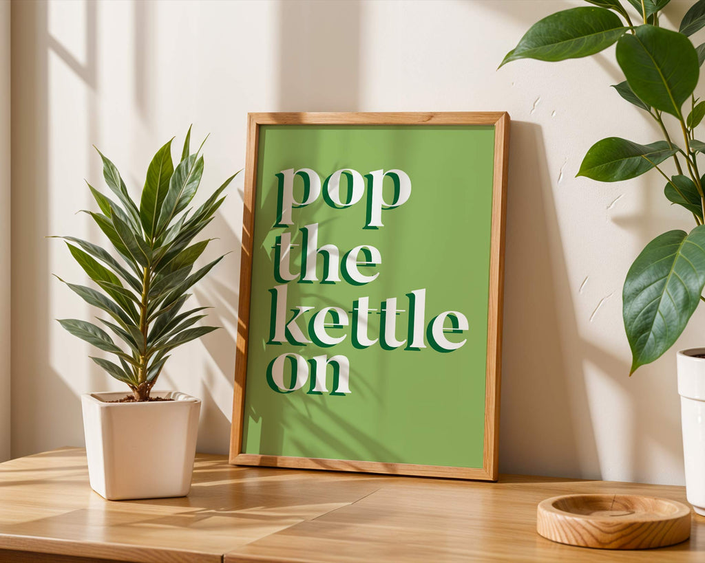 Framed artwork with 'pop the kettle on' text on a wooden table with plants.