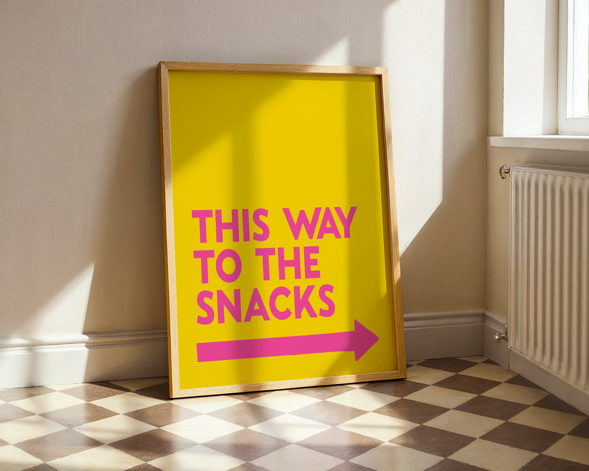 Framed poster with 'This way to the snacks' text on a yellow background in a room with sunlight.