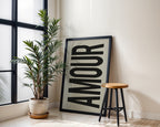 Framed artwork with the word 'AMOUR' next to a potted plant and stool in a room with large windows.