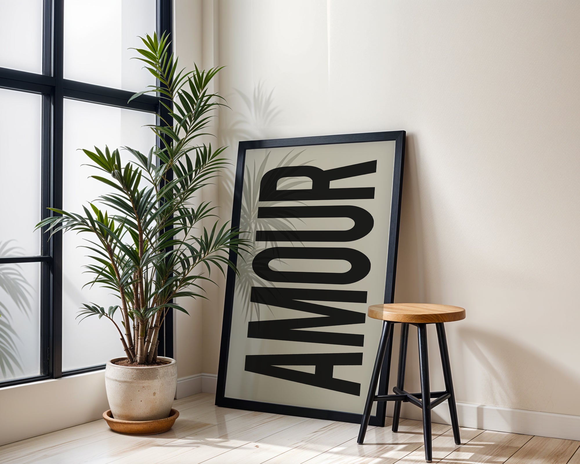 Framed artwork with the word 'AMOUR' next to a potted plant and stool in a room with large windows.