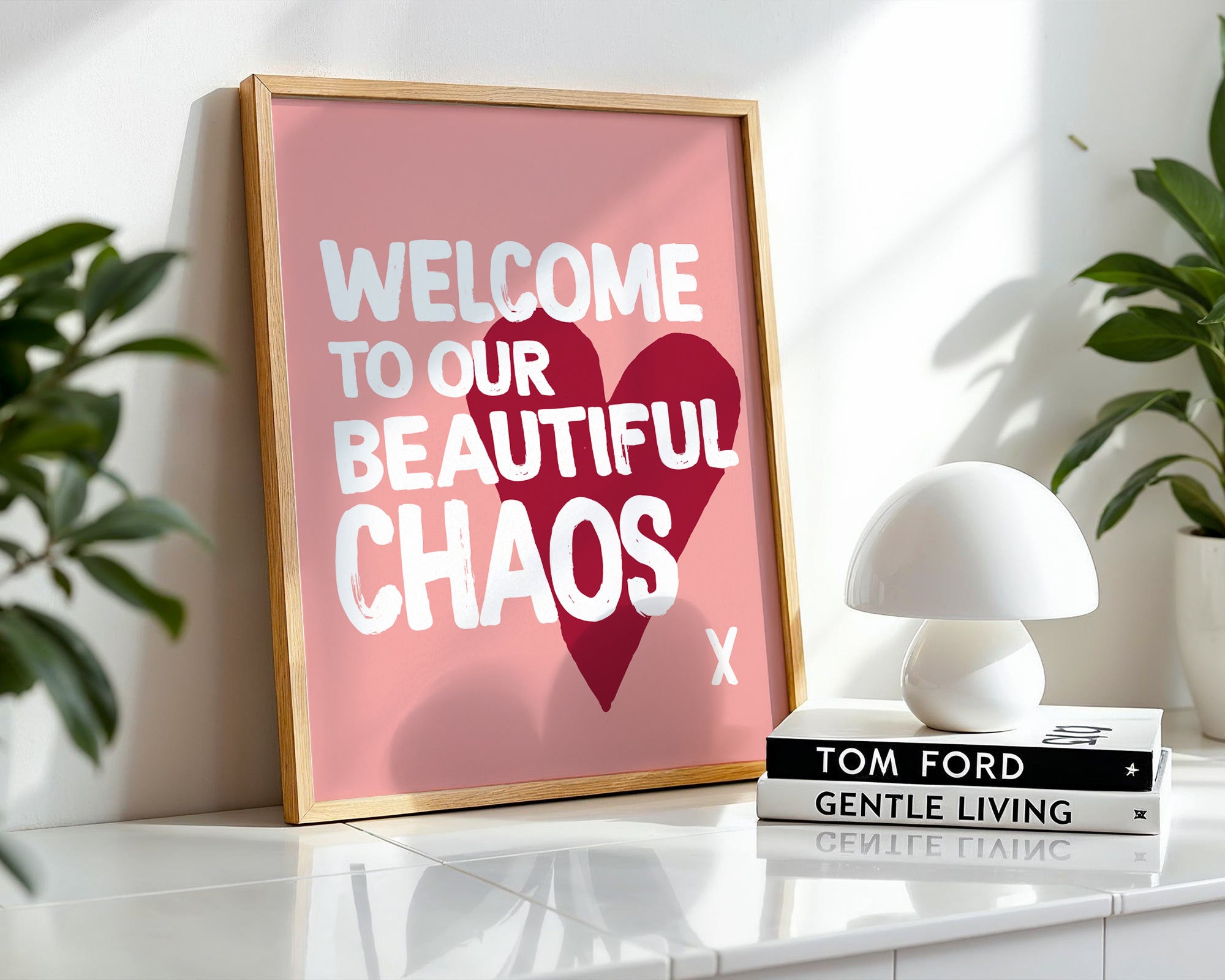 Framed poster with 'Welcome to our beautiful chaos' text on a shelf with books and a lamp.