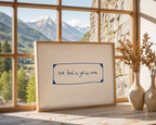 Framed artwork with 'the best is yet to come' quote in a room with large windows showing mountains.