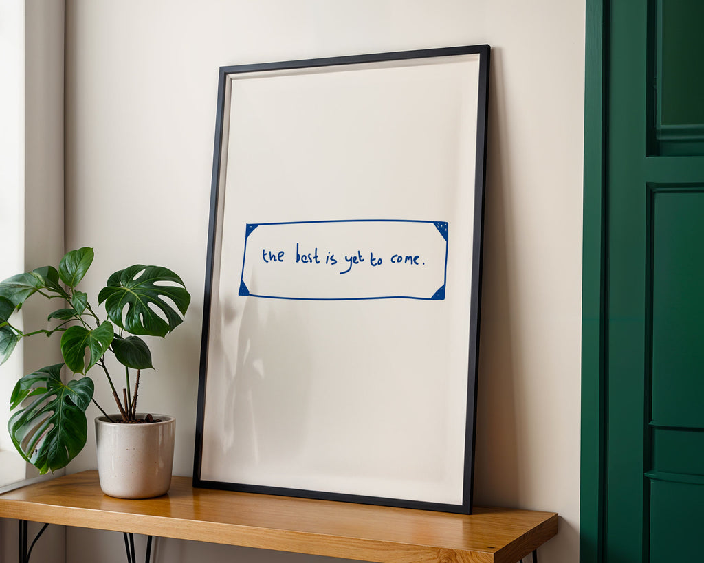 Framed print with text on a wall next to a plant and wooden shelf