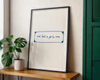 Framed print with text on a wall next to a plant and wooden shelf
