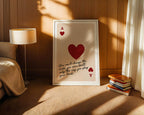 Framed artwork with a red heart and text in a cozy room with a lamp and books.