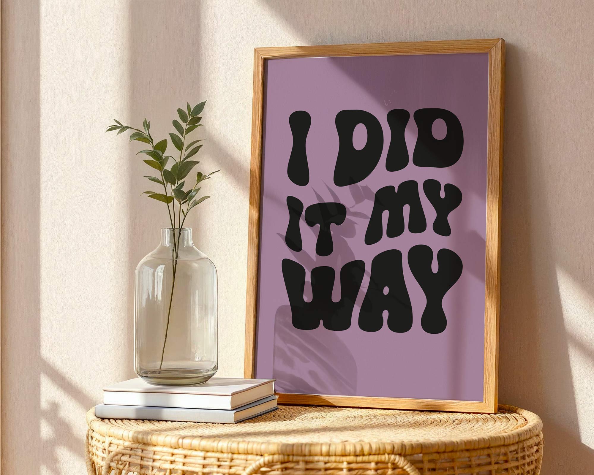 Framed poster with 'I DID IT MY WAY' text on a purple background, placed on a wicker table with a vase and books.