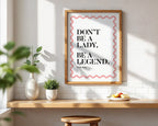 Framed quote 'Don't be a lady, be a legend' on a wall with a wooden shelf and plants.