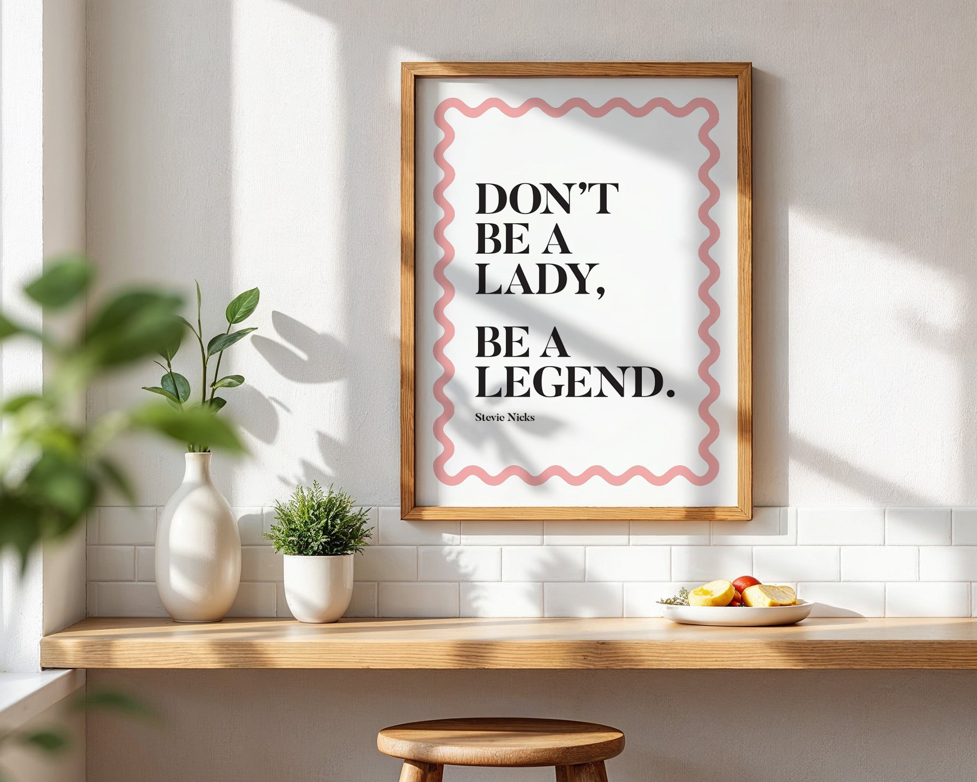 Framed quote 'Don't be a lady, be a legend' on a wall with a wooden shelf and plants.