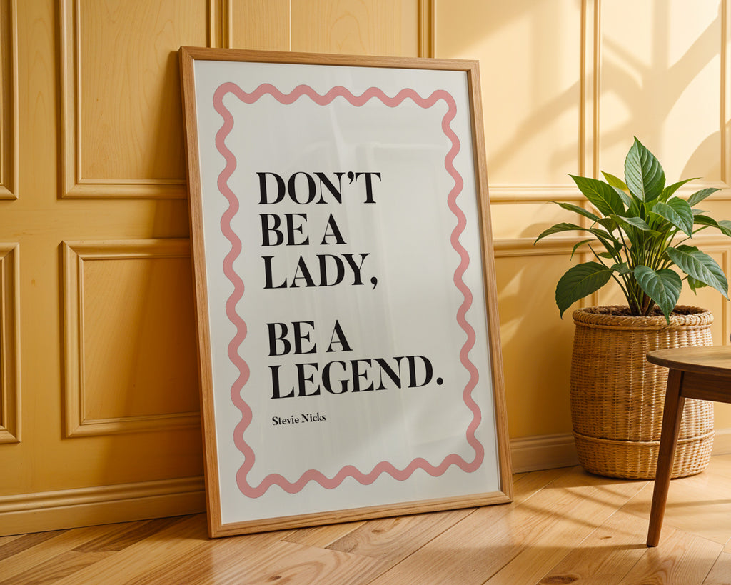 Framed poster with motivational quote 'Don't be a lady, be a legend' on a wooden floor with a plant in the background.