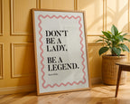 Framed poster with motivational quote 'Don't be a lady, be a legend' on a wooden floor with a plant in the background.