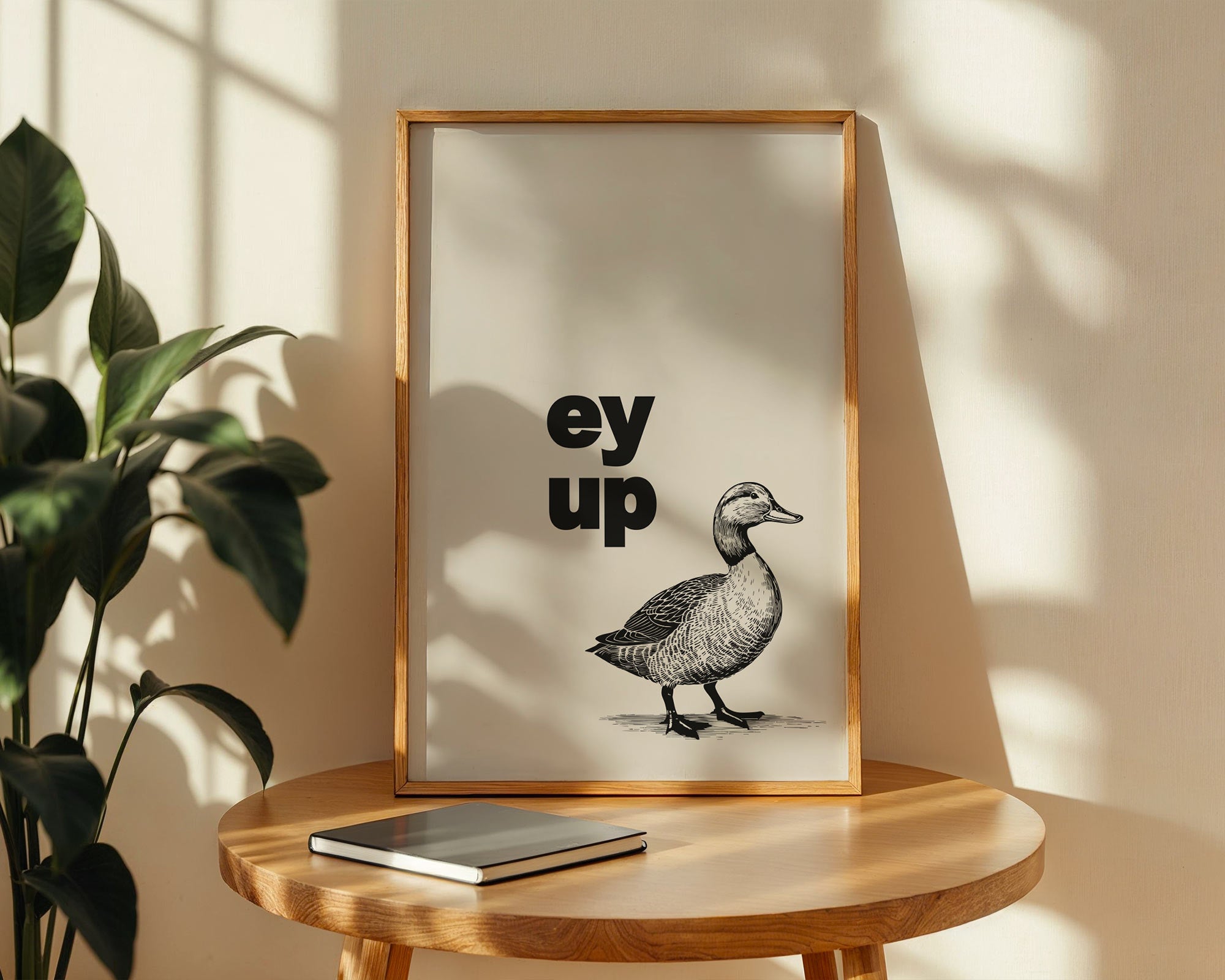 Framed artwork with a duck illustration and text on a wooden table with a plant in the background