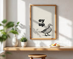 Framed artwork with a duck illustration and 'ey up' text on a wall with sunlight casting shadows.