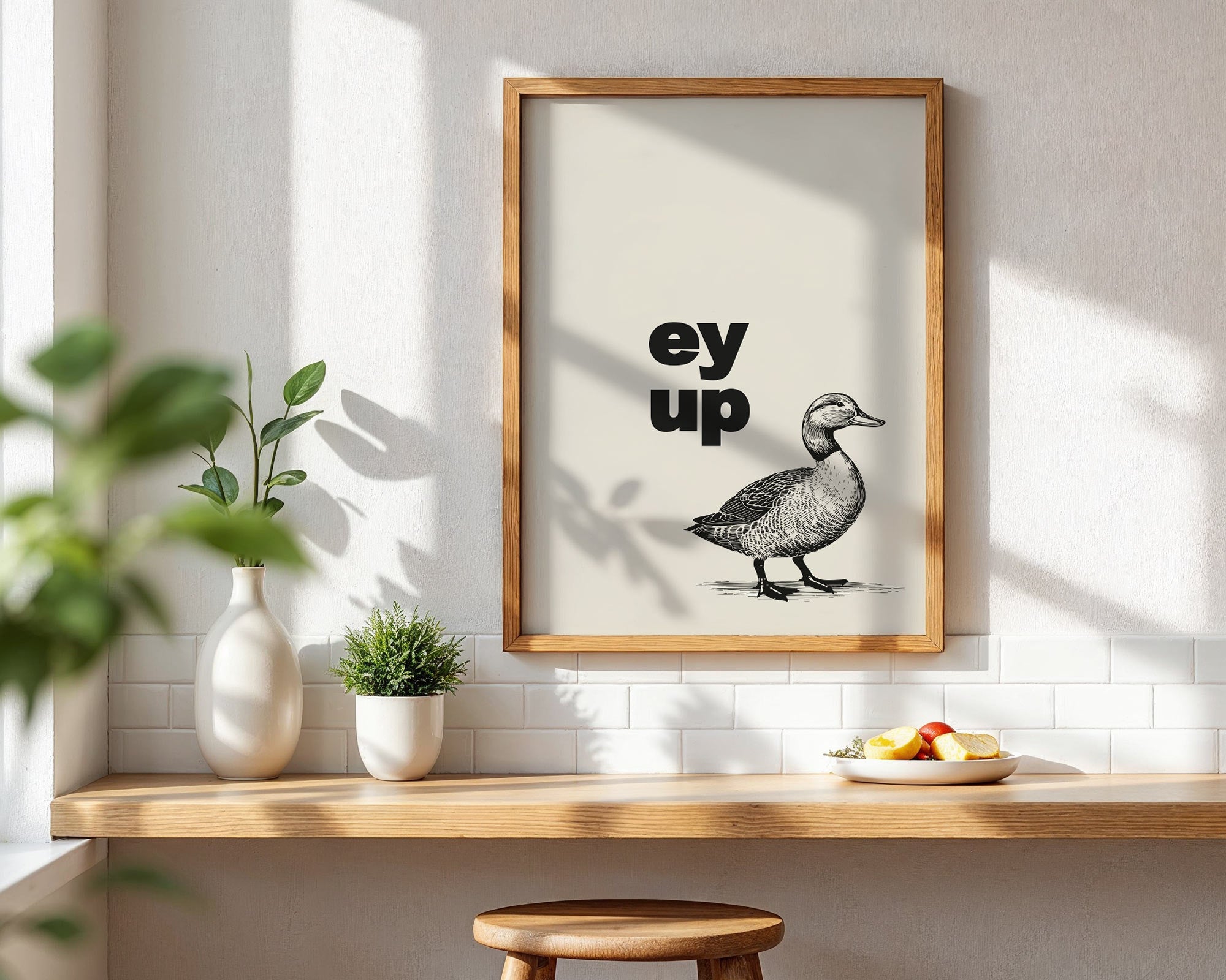Framed artwork with a duck illustration and 'ey up' text on a wall with sunlight casting shadows.