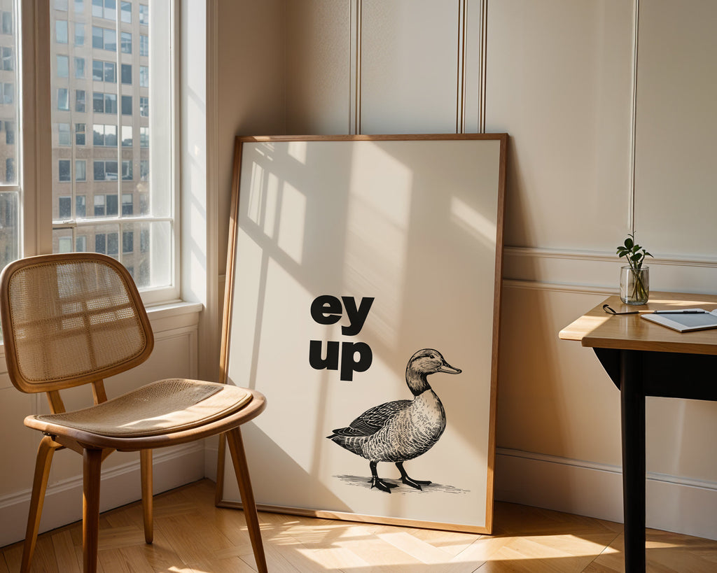 Framed artwork with a duck illustration and text in a room with a chair and table.