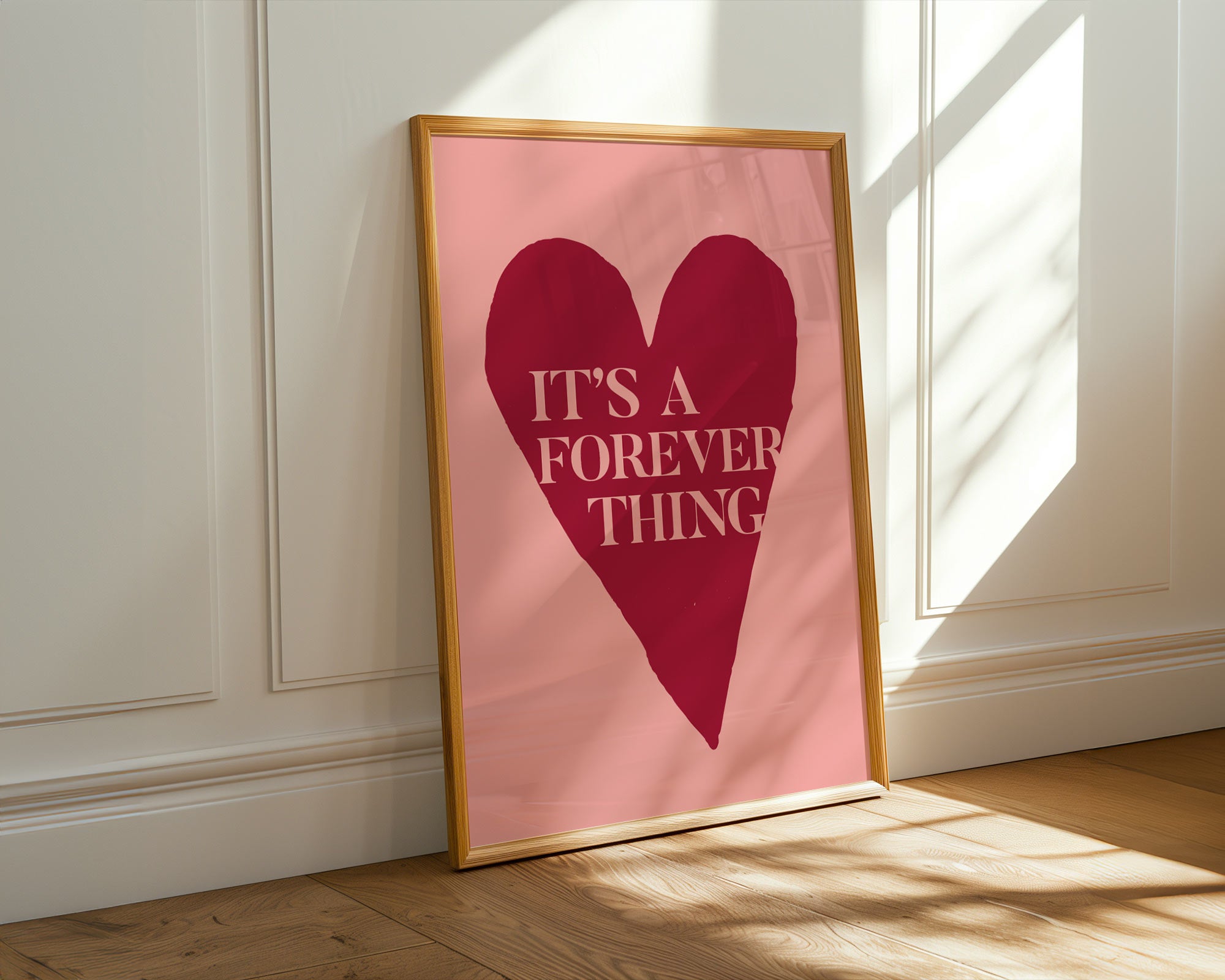 Framed poster with a red heart and text on a pink background, leaning against a white wall.