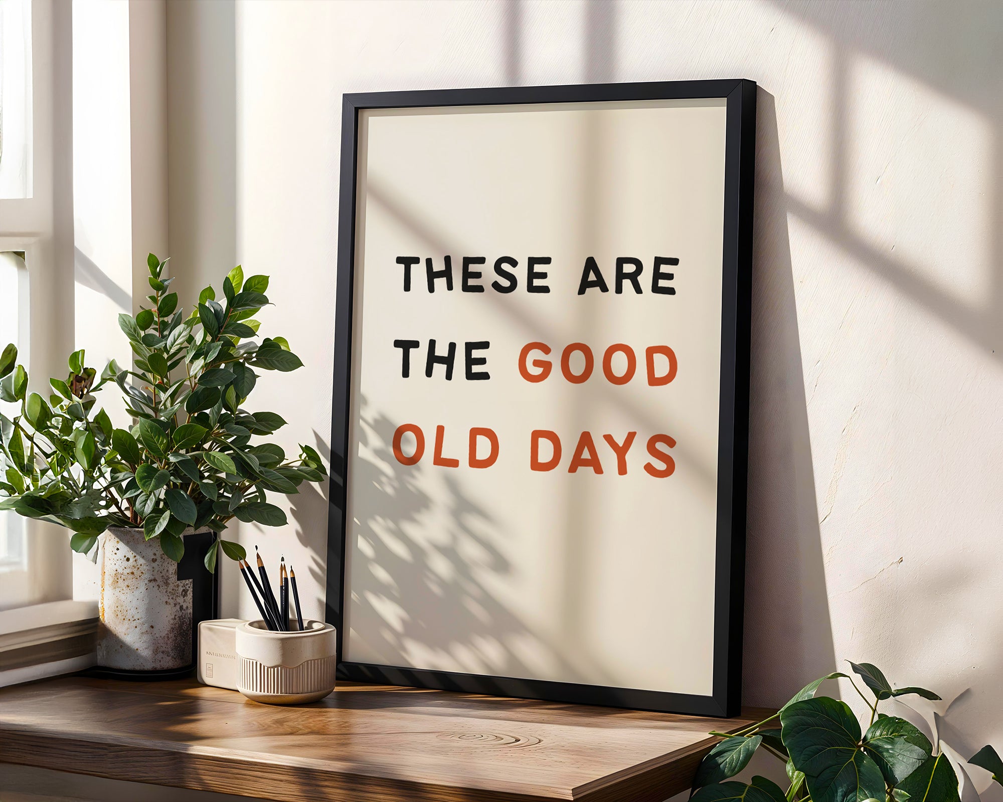 Framed poster with 'These are the good old days' text on a wall with plants and a window.
