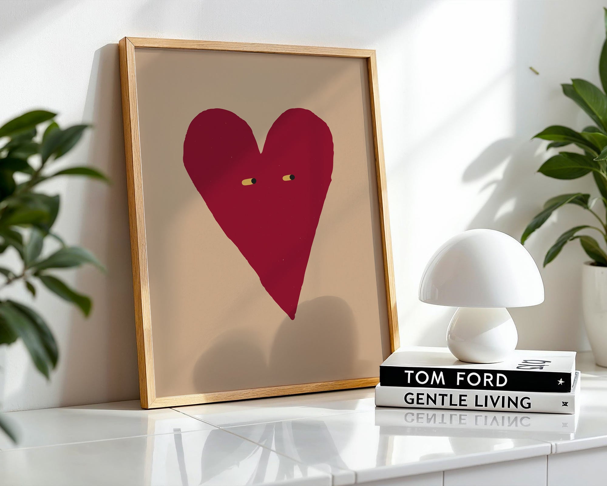 Framed artwork with a red heart on a beige background, placed on a white surface with plants in the background.