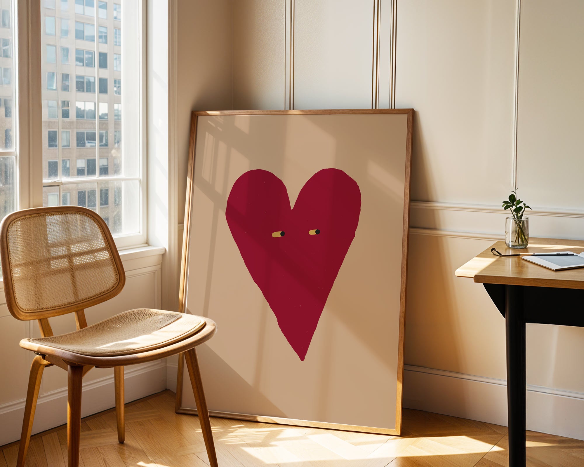 Framed artwork with a red heart on a beige background in a room with a chair and table.