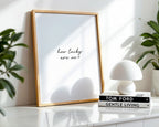 Framed print with 'how lucky are we?' text on a shelf with books and a lamp.