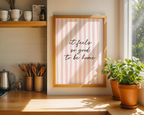 Framed print with 'it feels so good to be home' text in a kitchen setting with plants and shelves.