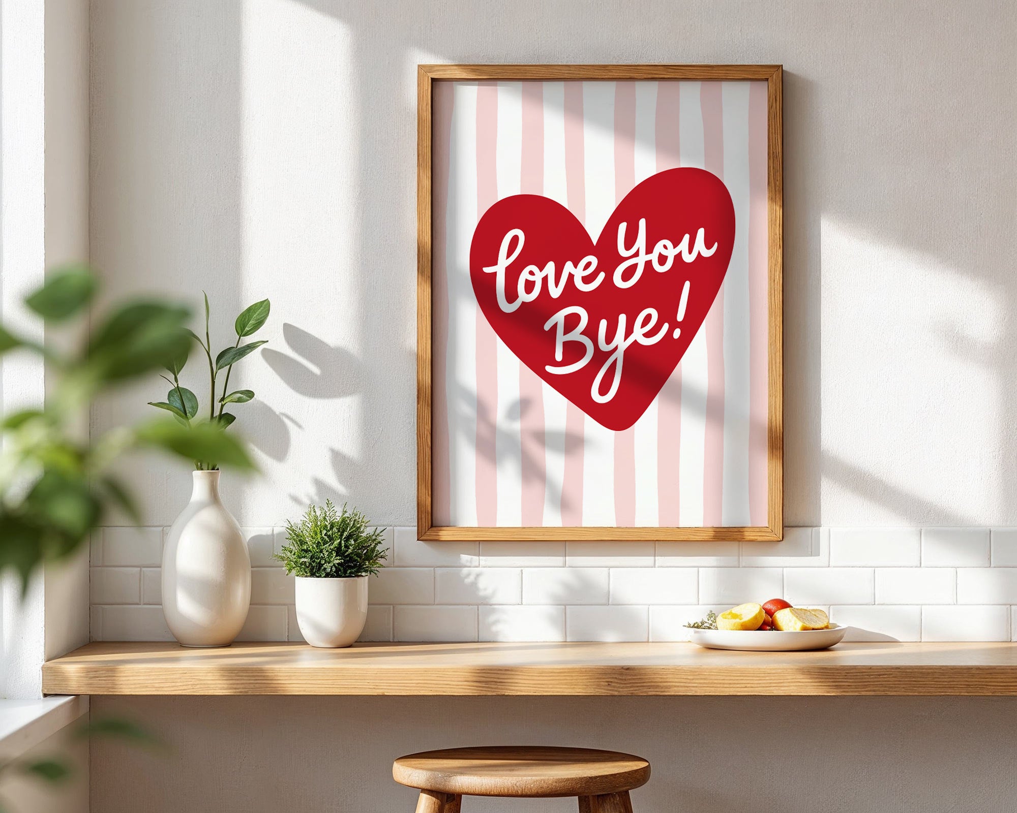 Framed artwork with a red heart and 'Love You Bye!' text on a striped background, placed on a wooden surface with plants and a stool.