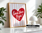Framed artwork with a red heart and 'Love You Bye!' text on a striped background, placed on a surface with books and a lamp.