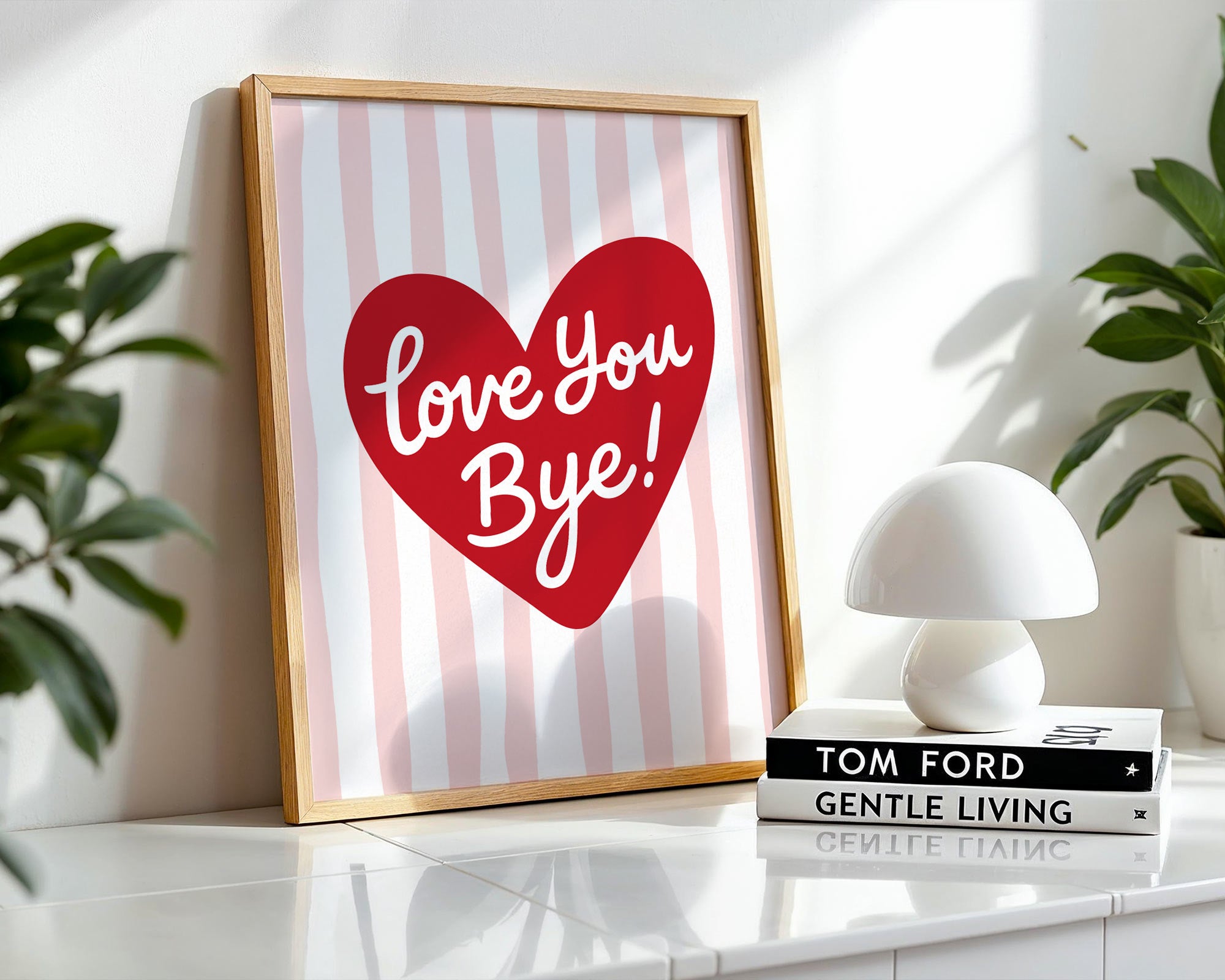 Framed artwork with a red heart and 'Love You Bye!' text on a striped background, placed on a surface with books and a lamp.