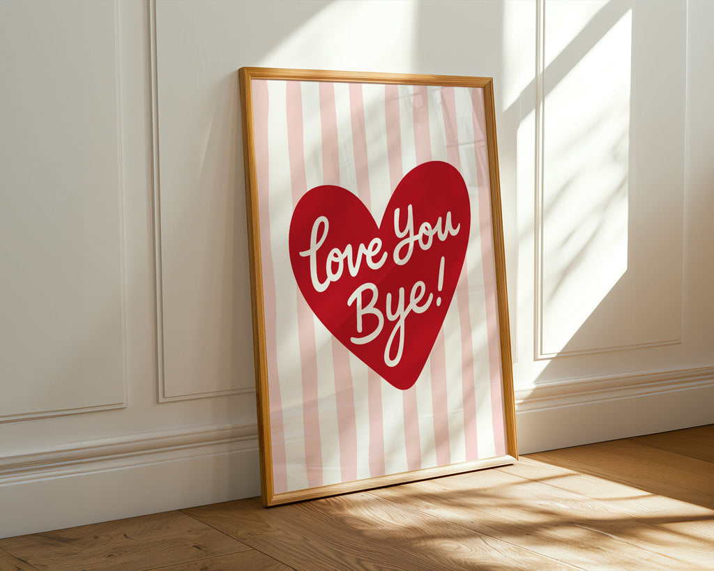Framed artwork with a red heart and 'Love You Bye!' text on a striped background, leaning against a white wall.