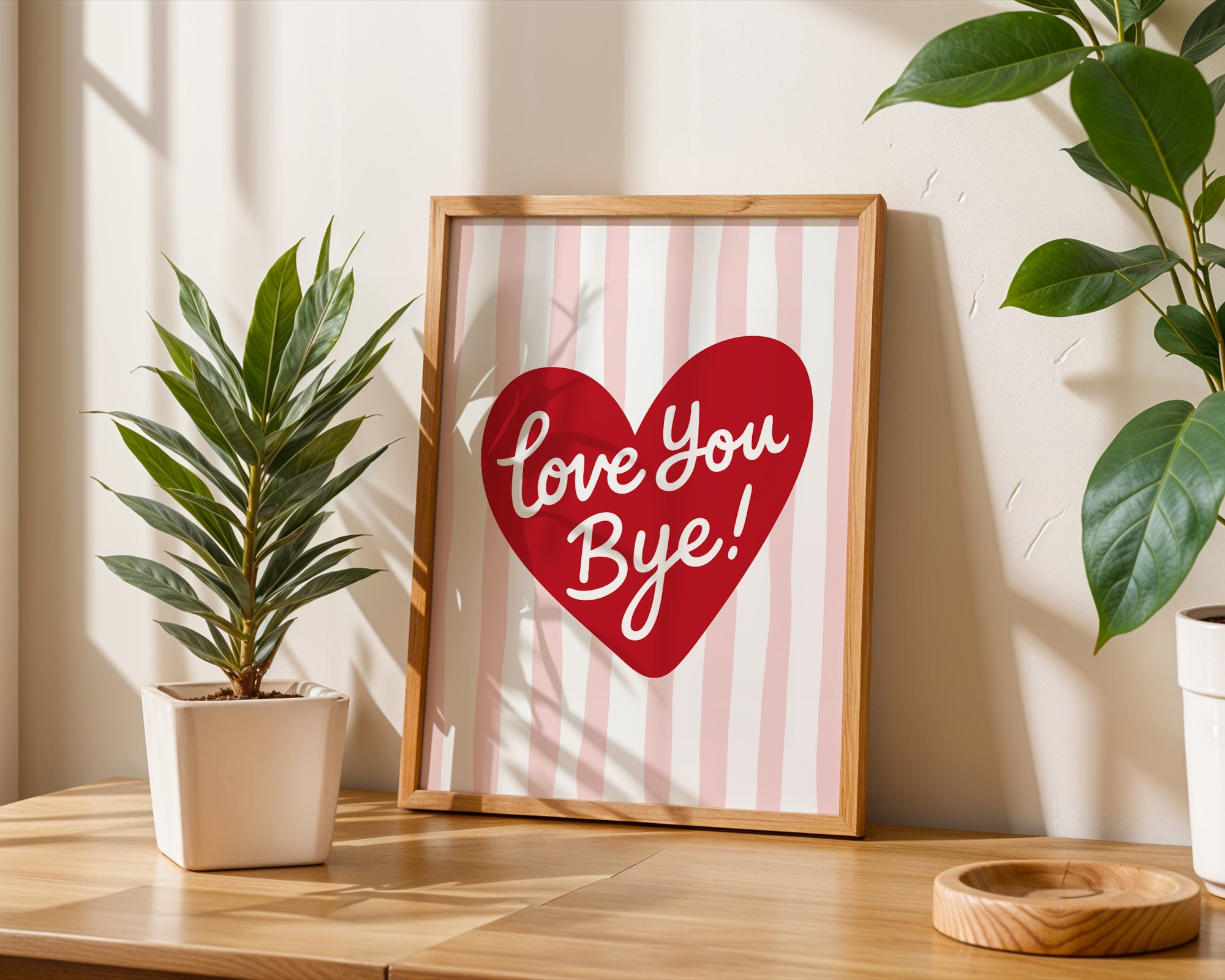 Framed artwork with 'Love You Bye!' on a striped background, placed on a wooden surface with plants.