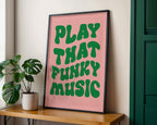 Framed poster with 'Play That Funky Music' text on a pink background, placed on a wooden shelf with a plant beside it.