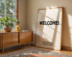 Room interior with a wooden sideboard, plant, and large framed sign with 'WELCOME! JUST KIDDING, PLEASE LEAVE' text.